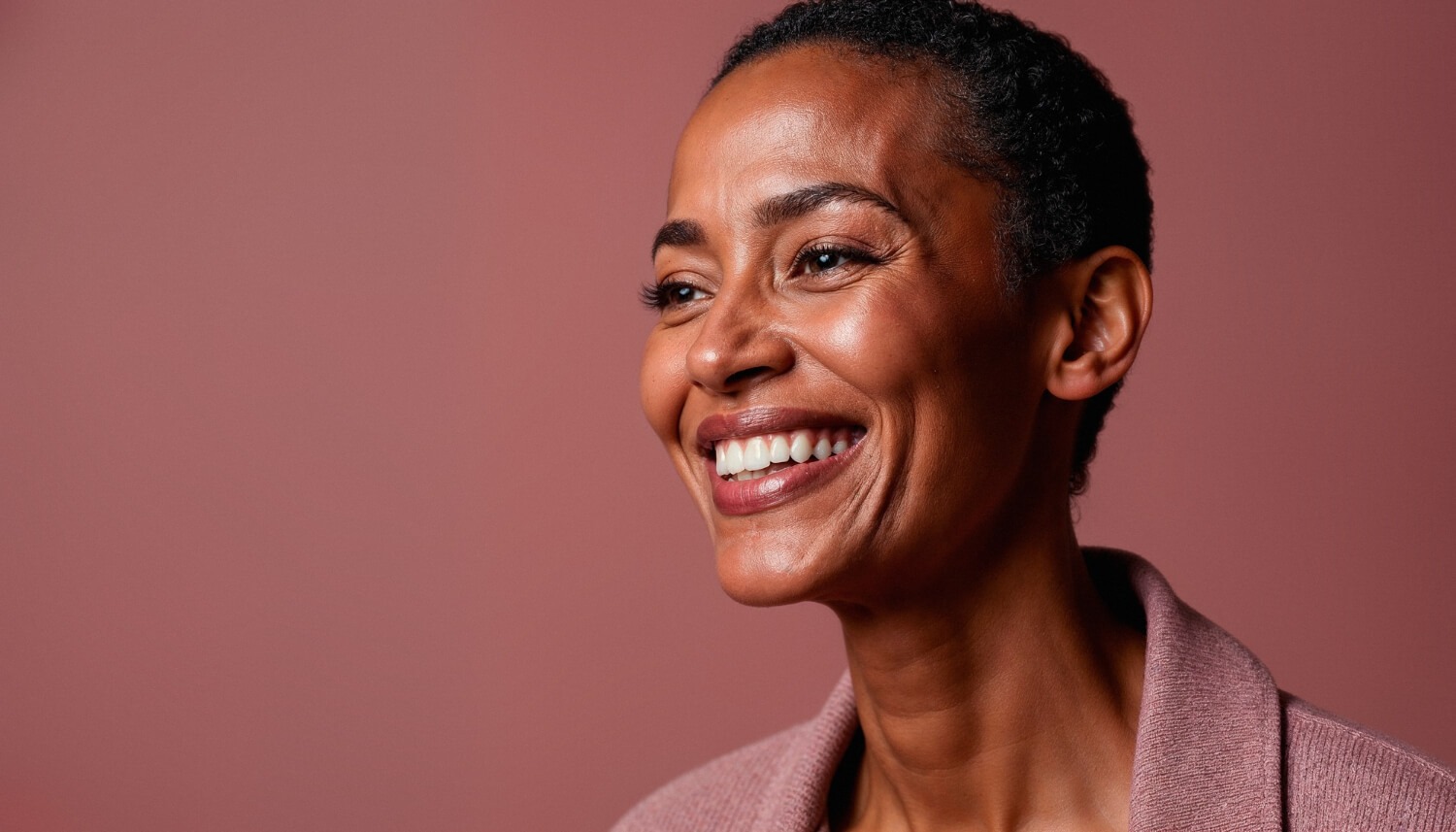 Mulher negra de cabelo curto sorrindo largamente para a direita em fundo rosado, exibindo dentes alinhados após tratamento com implantes dentários na Tijuca.